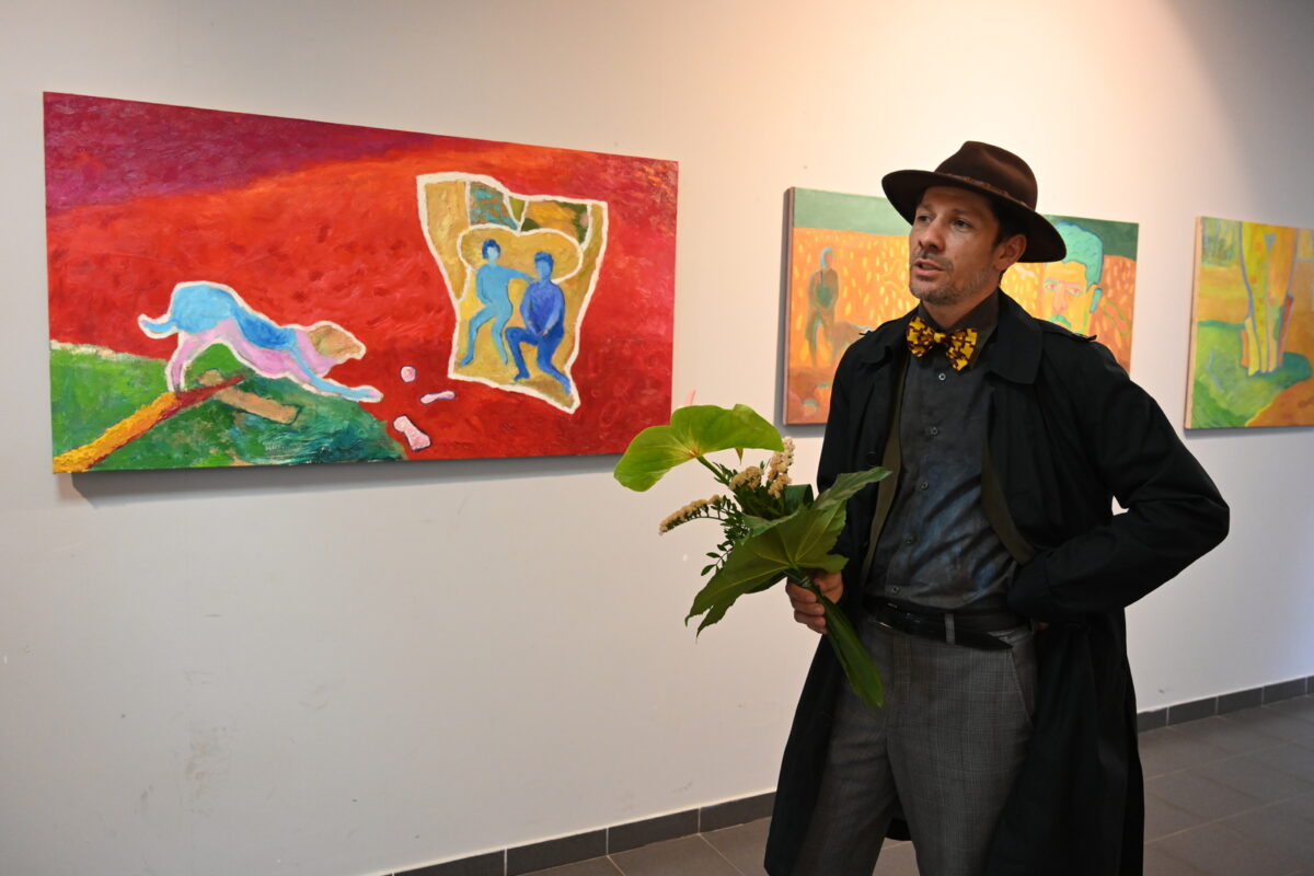 Artysta w kapeluszu z kwiatami na tle swoich obrazów.